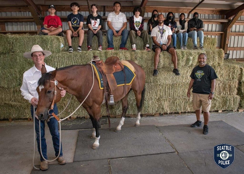A group of youth sitting on hay bales inside SPD's Mounted Patrol Unit barn with one horse